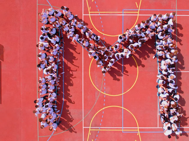 Students and staff forming the letter M on the red sports court, seen from the drone