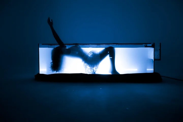 A figure in a glowing water tank in a dark room