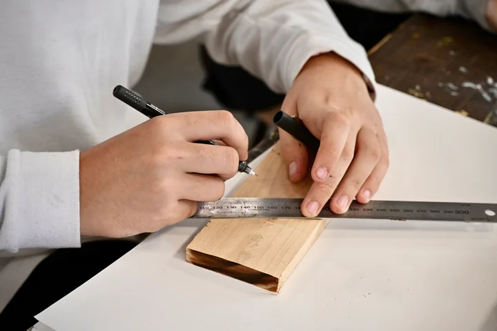 A student marking a line on wood with a pen and steel rule
