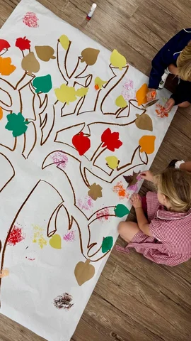 Primary pupils painting autumn leaves onto a giant paper tree on the classroom floor