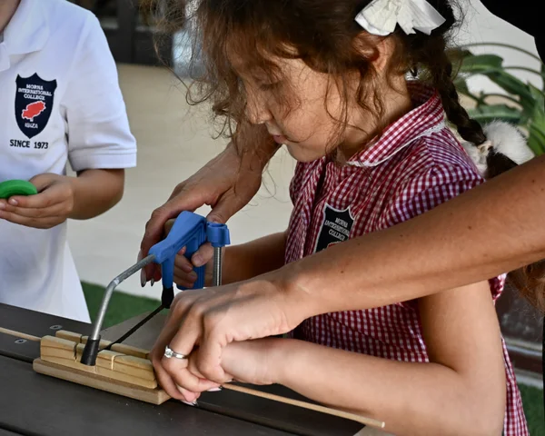 A girl in a gingham dress sawing a dowel with help from a teacher