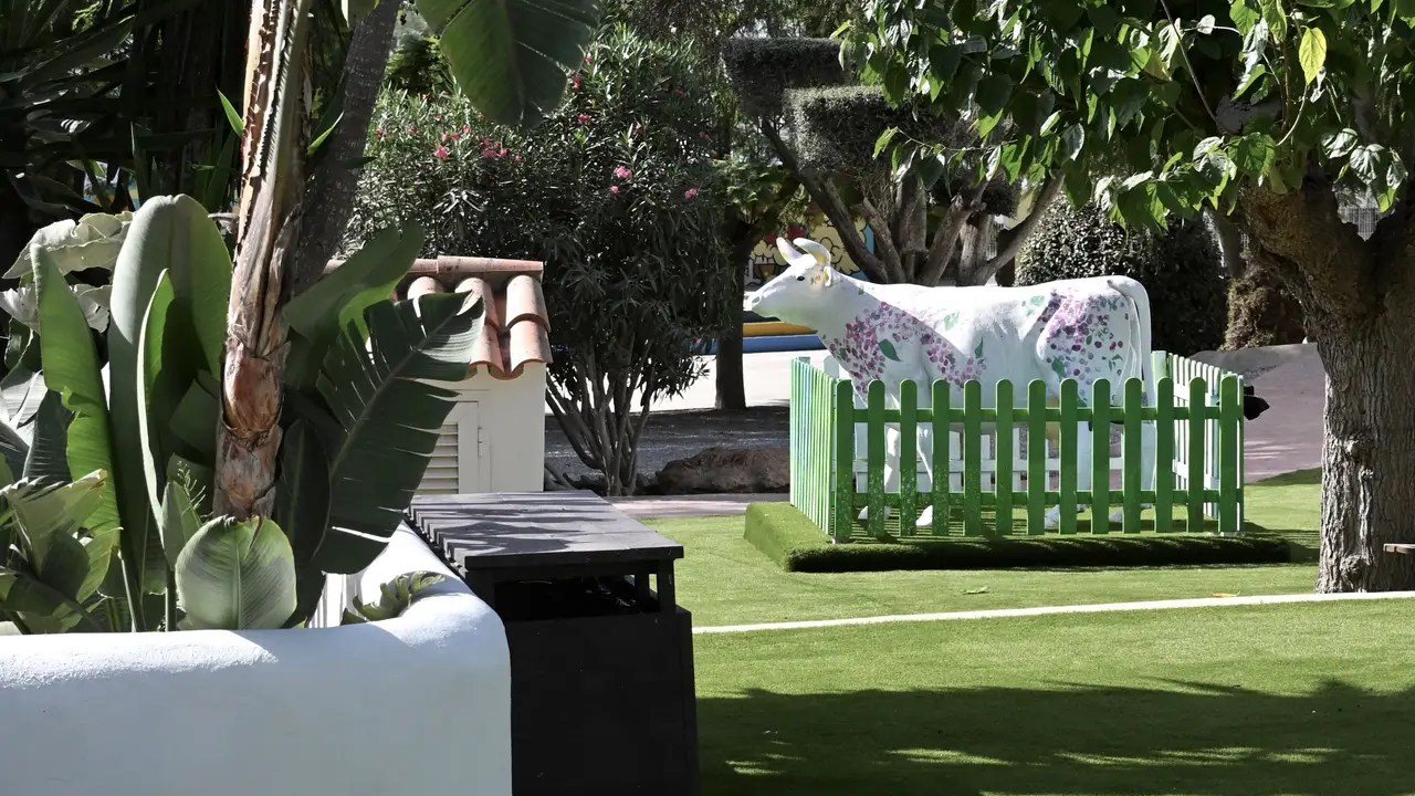 A painted cow sculpture behind a little green fence in the Morna garden, among palms and flowering shrubs