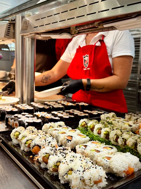 A kitchen team member in a red apron arranging trays of sushi rolls