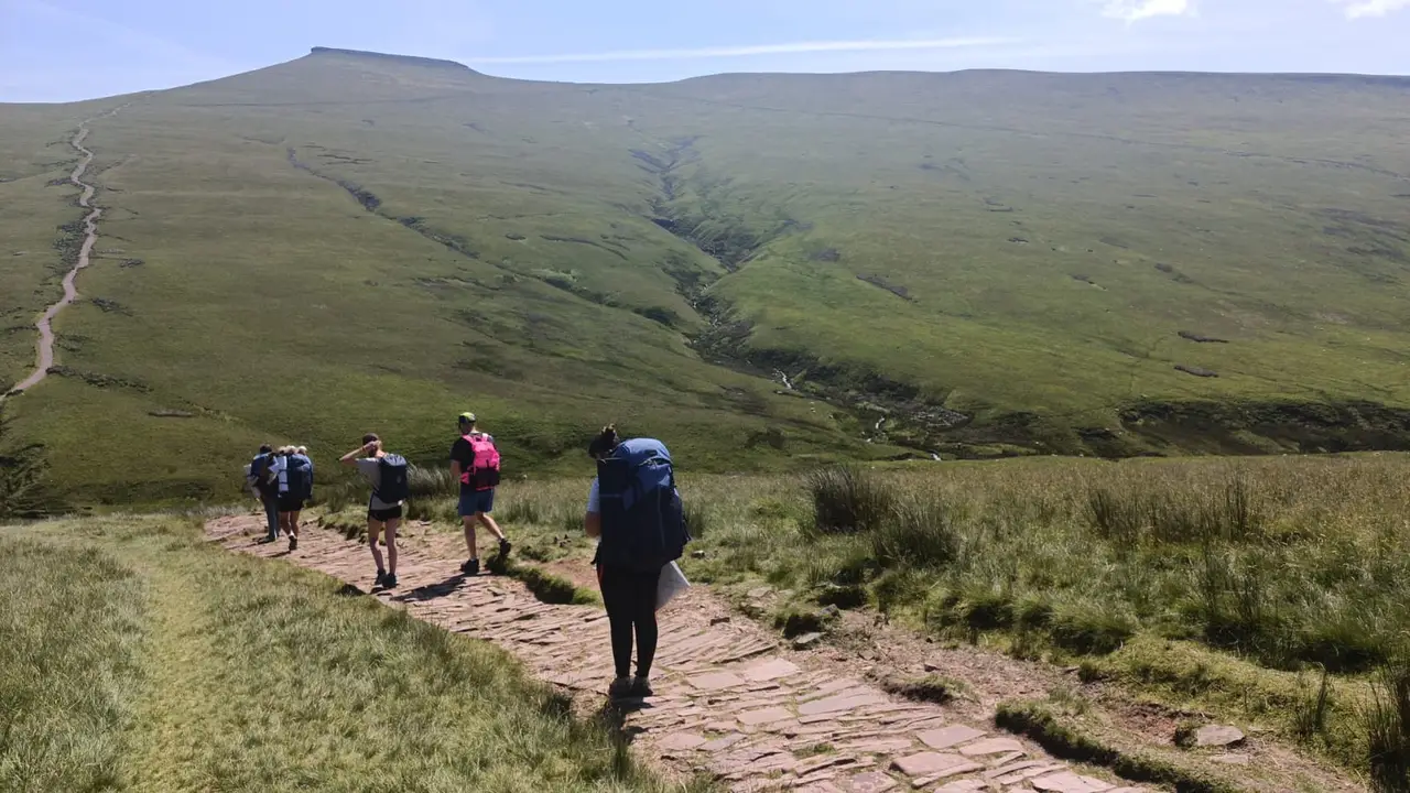 Students with rucksacks hiking up a stone path across open hills